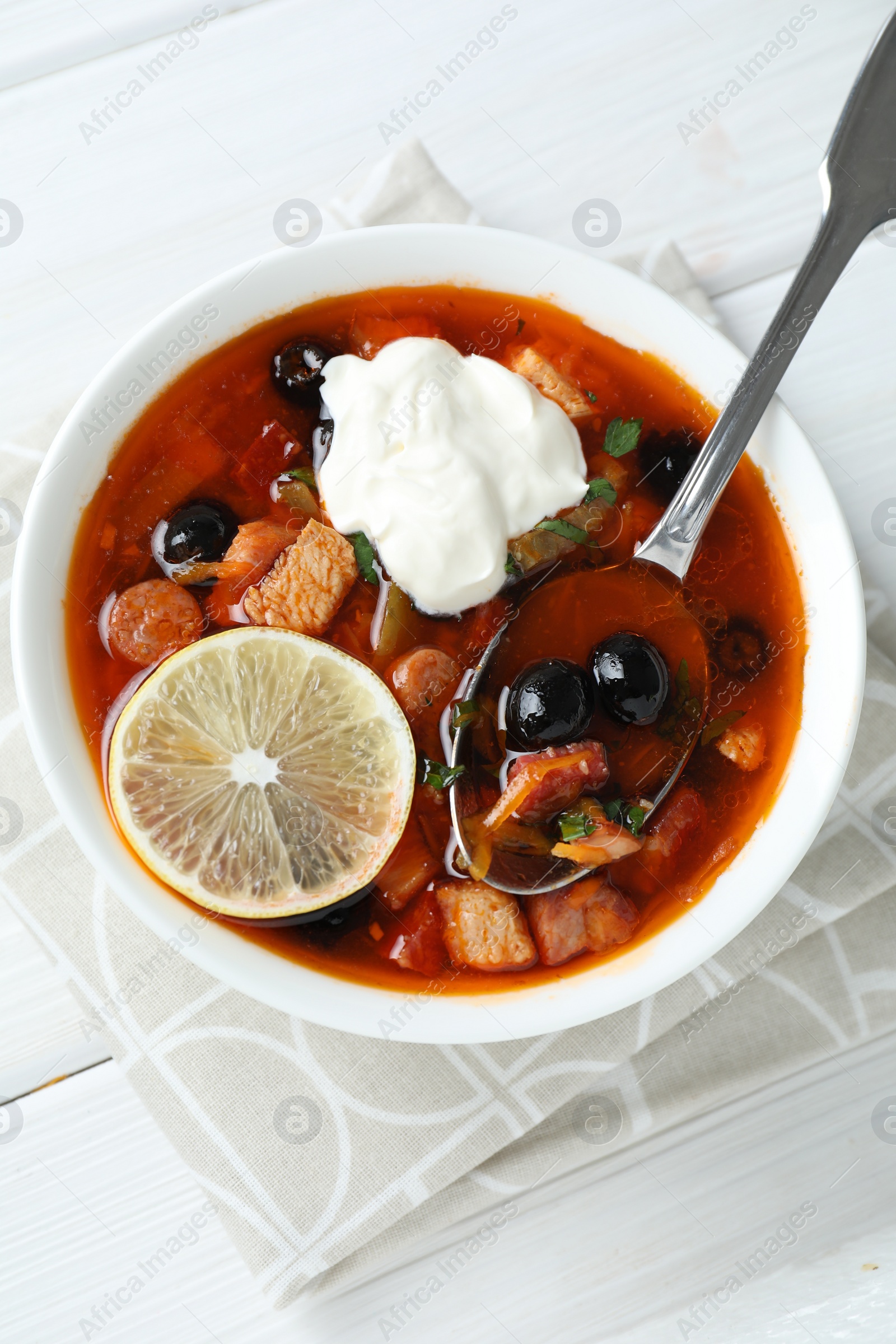 Photo of Meat solyanka soup with sausages, olives, vegetables and spoon on white wooden table, top view