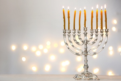 Photo of Silver menorah with burning candles against light grey background and blurred festive lights, space for text. Hanukkah celebration
