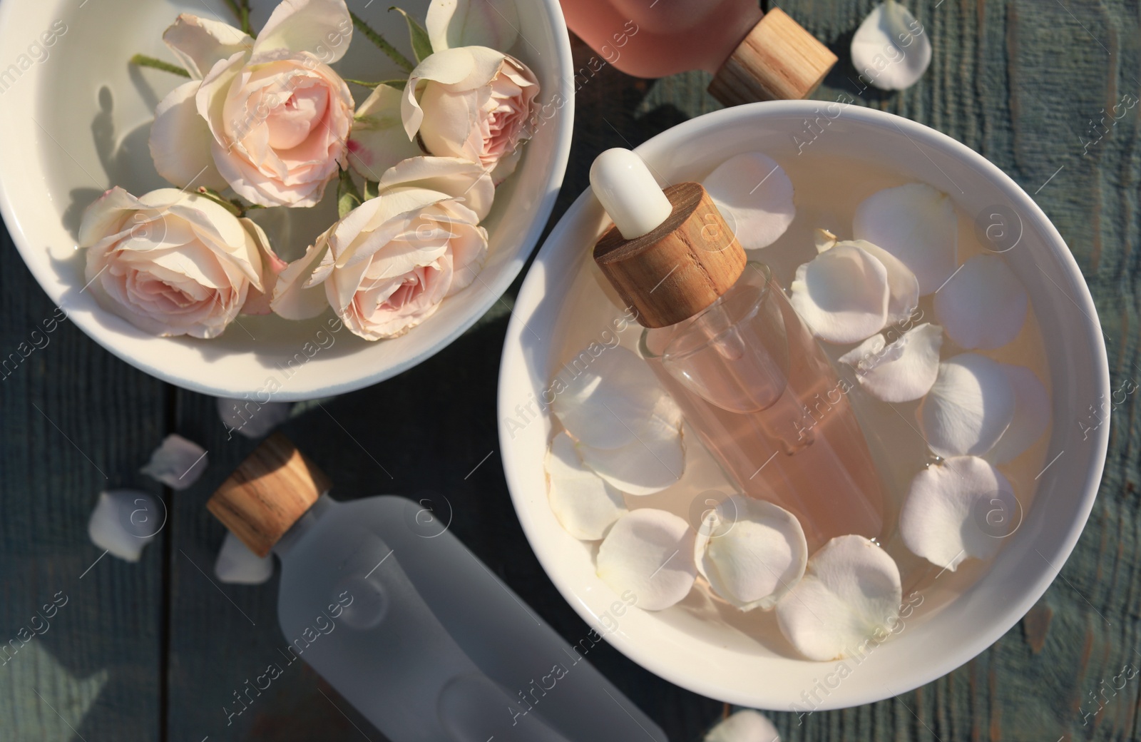 Photo of Bottles of rose essential oil and flowers on light blue wooden table, flat lay