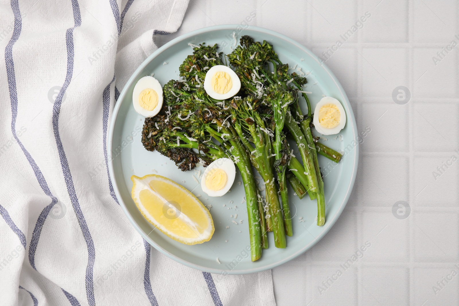 Photo of Tasty cooked broccolini with cheese, quail eggs and lemon on white tiled table, top view