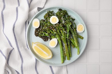 Tasty cooked broccolini with cheese, quail eggs and lemon on white tiled table, top view