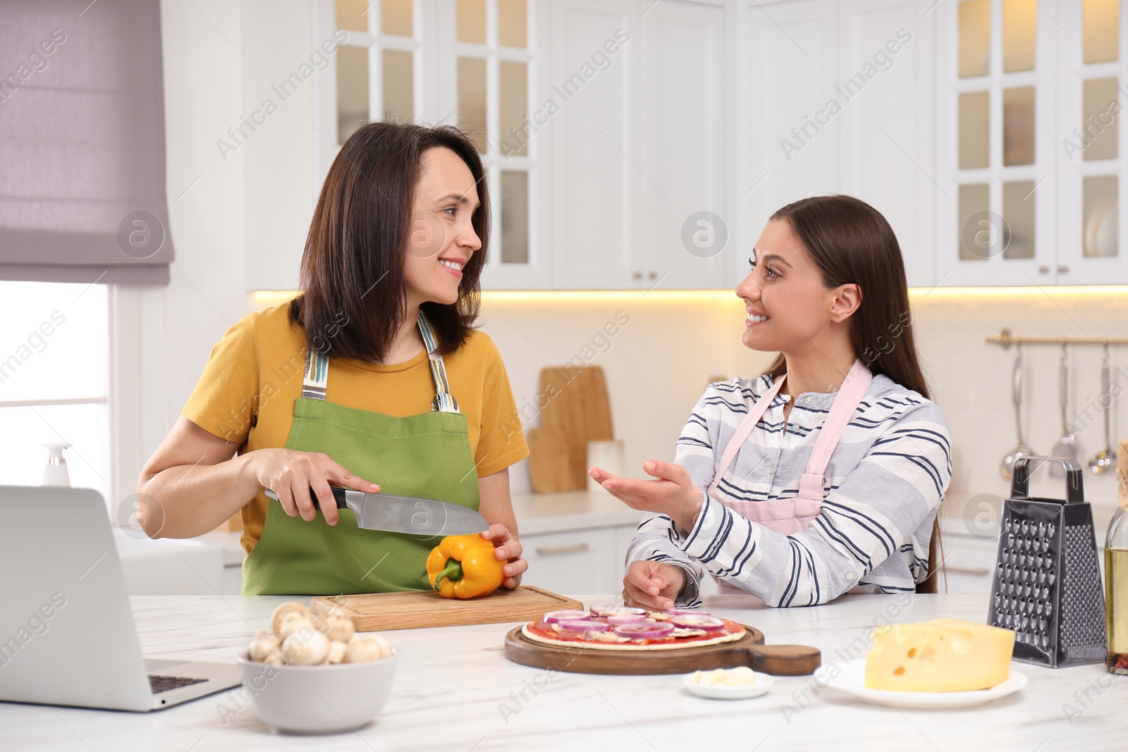 Photo of Mother with adult daughter making dinner while watching online cooking course via laptop in kitchen