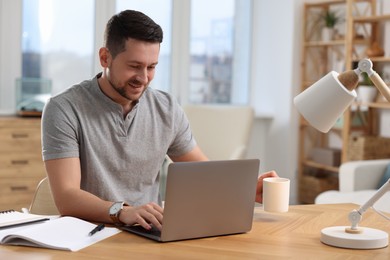 Happy man with cup of drink working on laptop at wooden desk in room