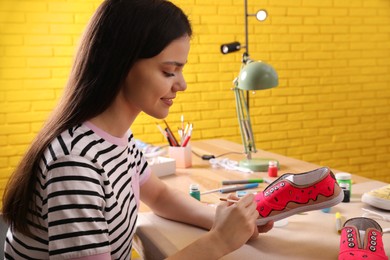 Photo of Woman painting on sneaker at wooden table indoors. Customized shoes