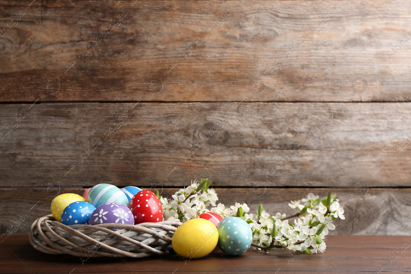 Photo of Painted Easter eggs in nest and blossoming branches on table against color background. Space for text