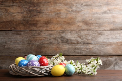 Photo of Painted Easter eggs in nest and blossoming branches on table against color background. Space for text