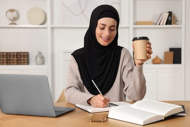 Muslim woman with cup of coffee writing notes near laptop at wooden table in room