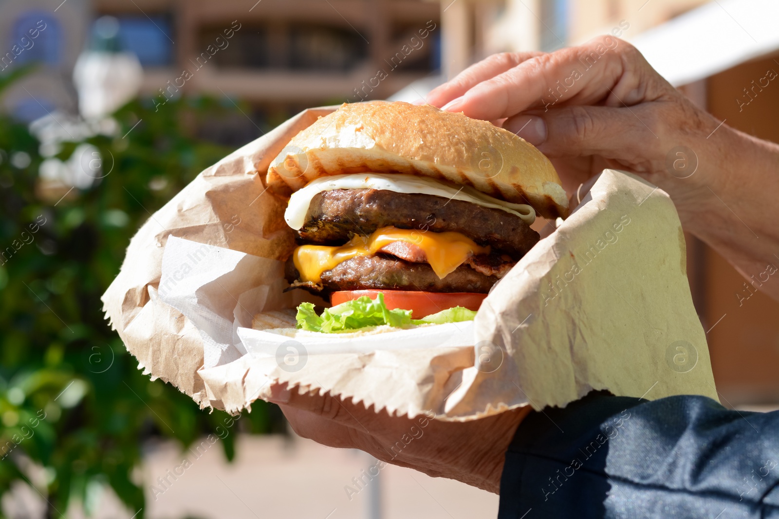 Photo of Woman holding delicious burger in paper wrap outdoors, closeup