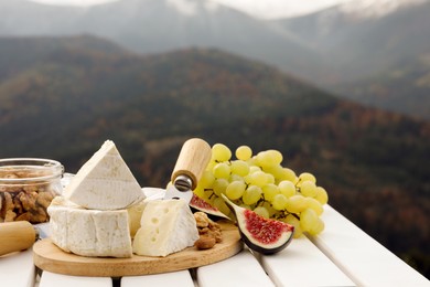 Delicious cheese, nuts and fruits on white wooden table against mountain landscape