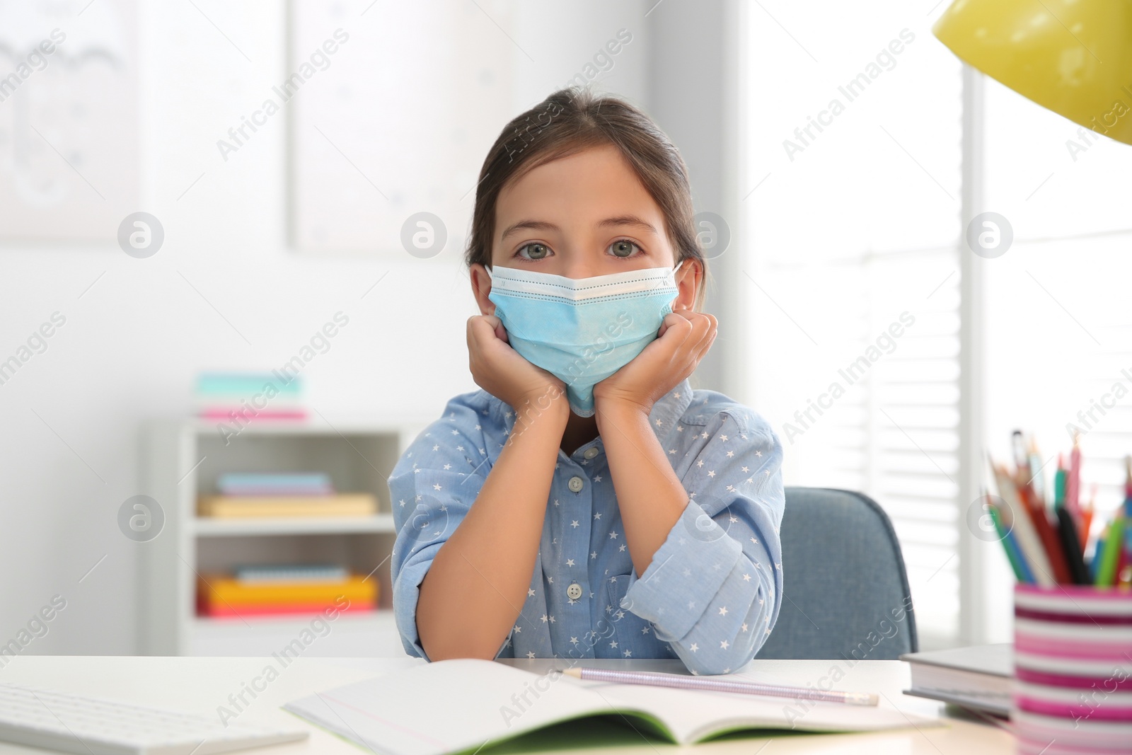 Photo of Distance learning, online studying at home due to Covid-19 pandemic. Girl in protective mask doing homework in room