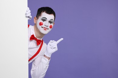 Photo of Funny mime artist peeking out of blank poster and pointing at something on purple background. Space for text