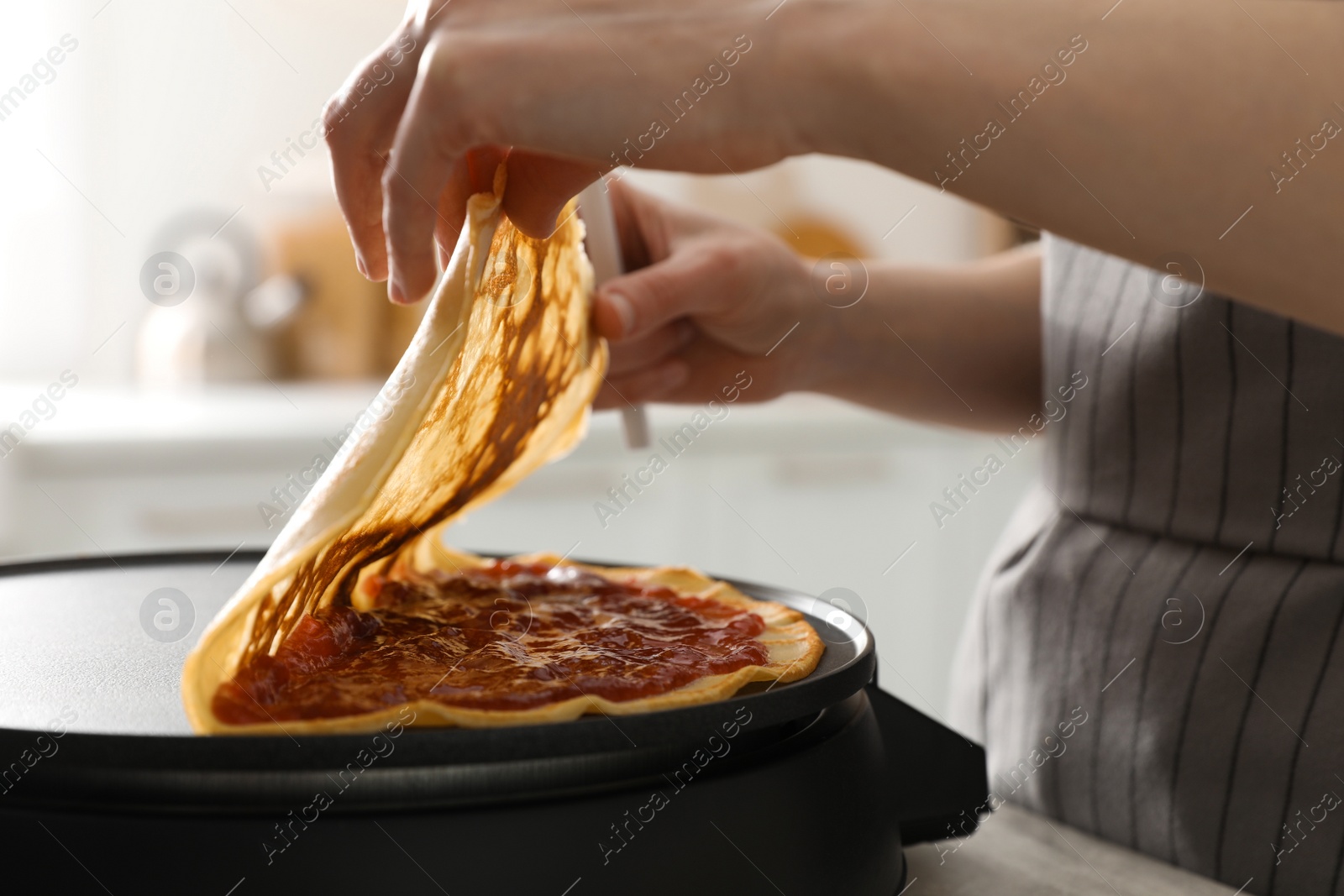 Photo of Woman cooking delicious crepe with jam on electric pancake maker in kitchen, closeup