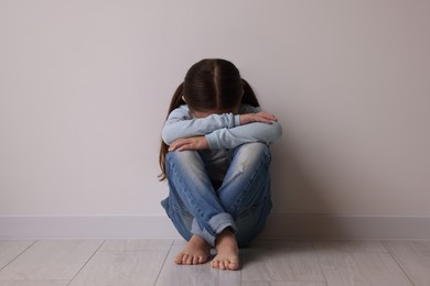 Photo of Child abuse. Upset little girl sitting on floor near light wall indoors