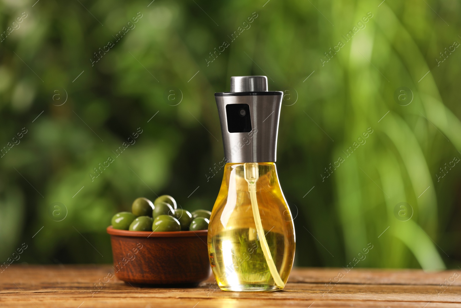 Photo of Spray bottle with cooking oil and olives on wooden table against blurred background, closeup