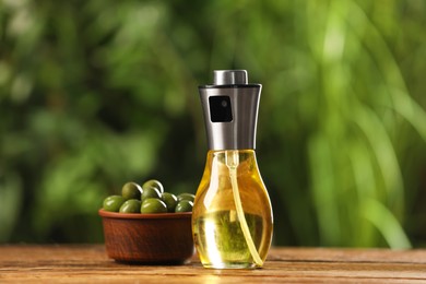 Spray bottle with cooking oil and olives on wooden table against blurred background, closeup