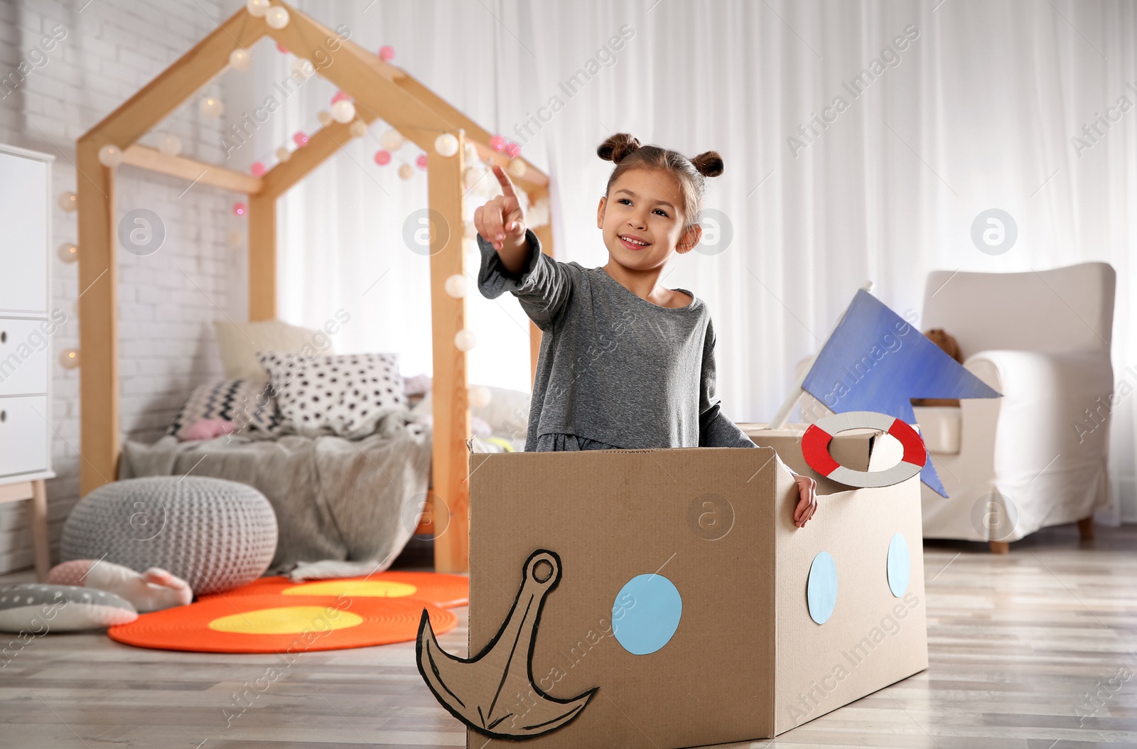 Photo of Cute little girl playing with cardboard boat in bedroom