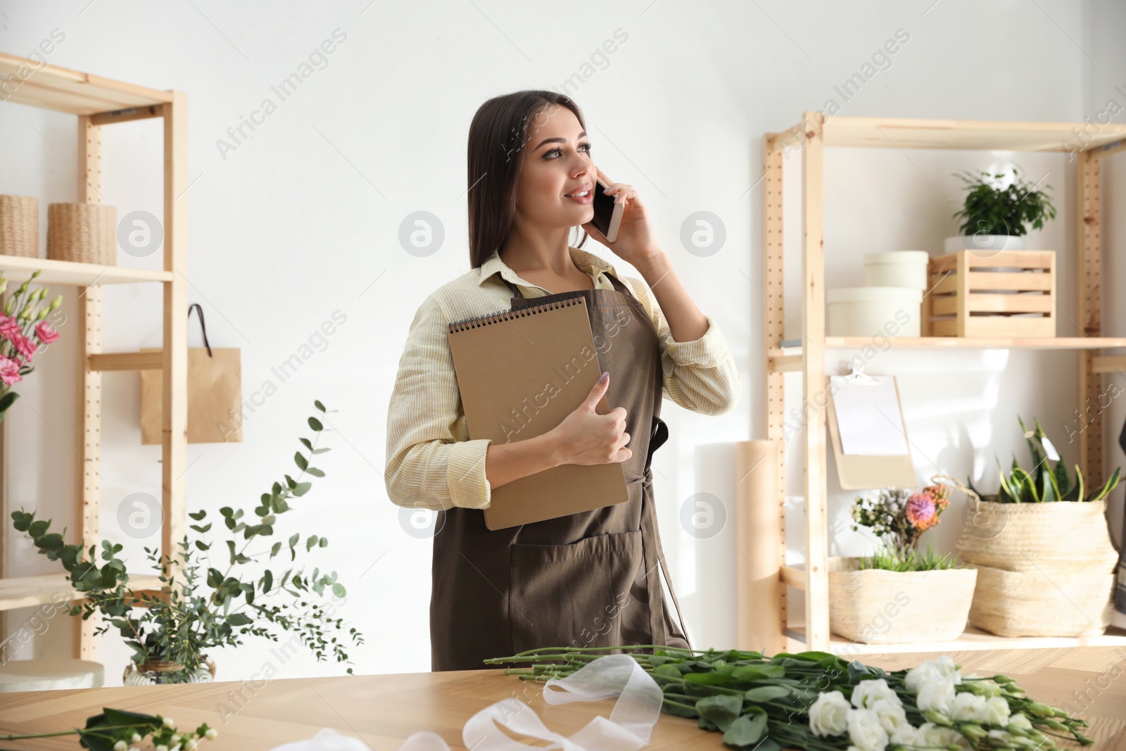 Photo of Florist with notebook talking on smartphone in workshop