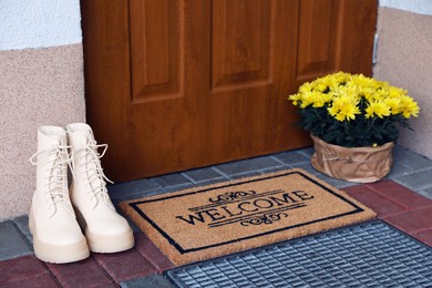 Photo of Door mat with word Welcome, stylish boots and beautiful flowers near entrance