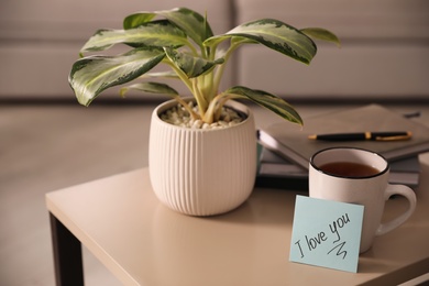 Paper note with handwritten words I Love You, cup of tea and houseplant on table indoors