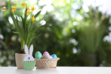 Photo of Easter bunny figure, dyed eggs and flowers on white wooden table against blurred green background. Space for text