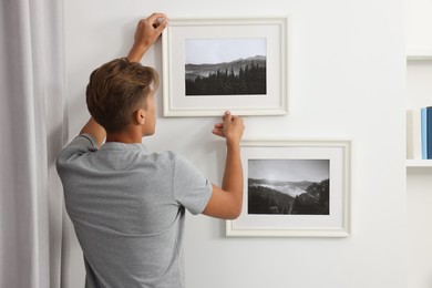 Photo of Young man hanging picture frames on white wall indoors, back view