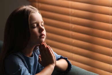 Cute little girl praying near window. Space for text
