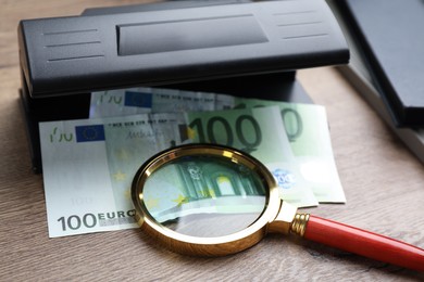 Modern currency detector with Euro banknotes and magnifying glass on wooden table. Money examination device