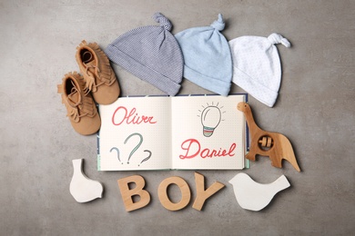 Photo of Notebook with written baby names, child's clothes and wooden letters on grey table, flat lay