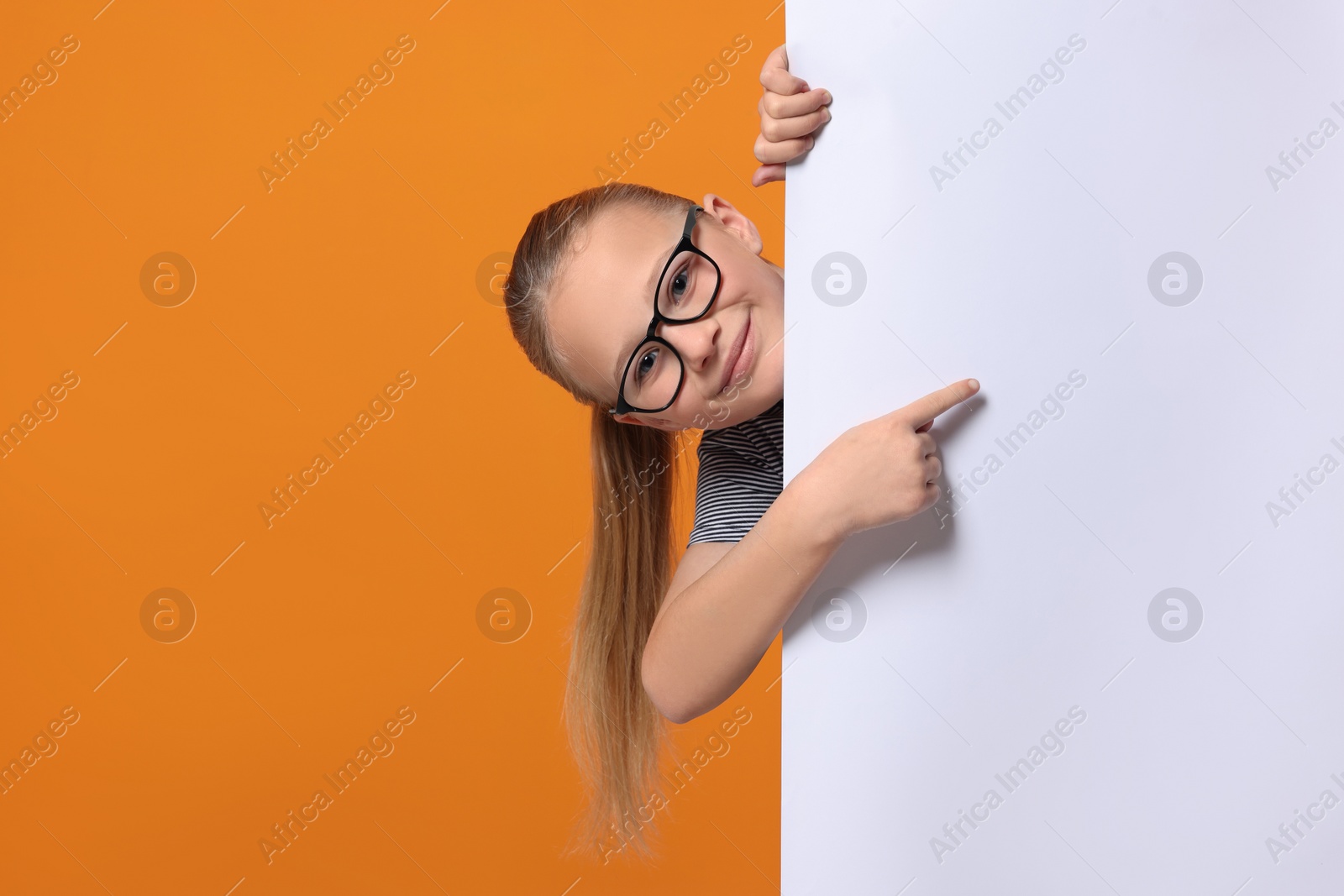 Photo of Cute girl looking out of placard and pointing on it against orange background. Space for text