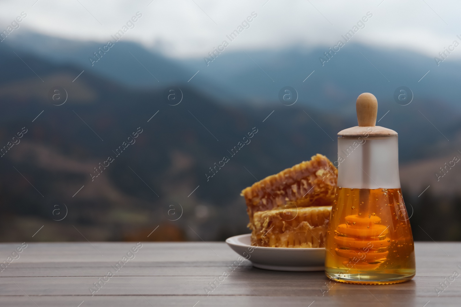 Photo of Fresh aromatic honey and combs on grey wooden table against mountain landscape. Space for text