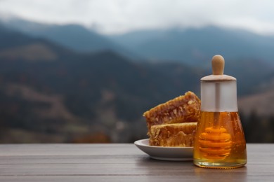 Fresh aromatic honey and combs on grey wooden table against mountain landscape. Space for text