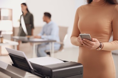 Photo of Employee with smartphone near new modern printer in office, closeup