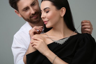 Man putting elegant necklace on beautiful woman against grey background, closeup