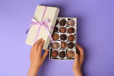 Photo of Child opening box of delicious chocolate candies on light purple background, top view