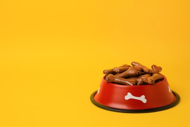 Photo of Red bowl with bone shaped dog cookies on yellow background, space for text