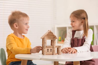 Photo of Little boy and girl playing with wooden house at white table indoors. Children's toys