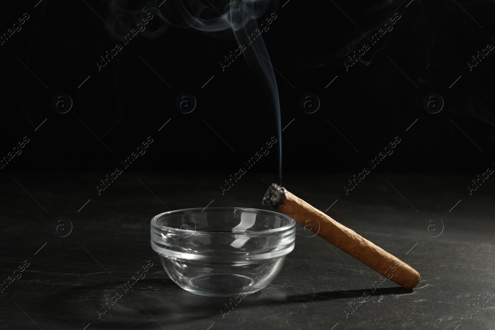 Photo of Smoldering cigar near glass ashtray on black table