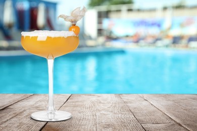 Tasty refreshing cocktail on wooden table near outdoor swimming pool at resort, space for text