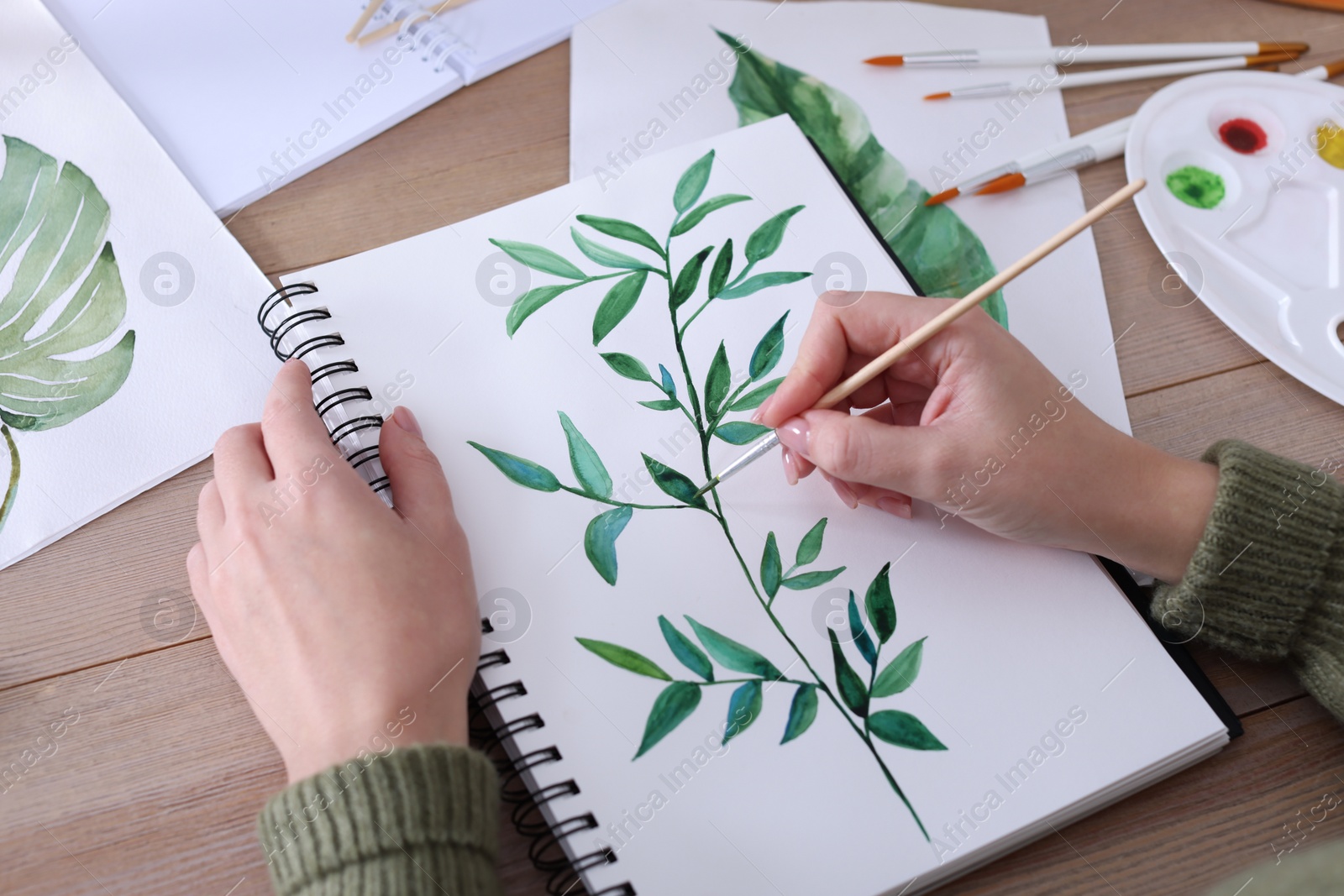 Photo of Woman painting green twig in sketchbook at wooden table, closeup