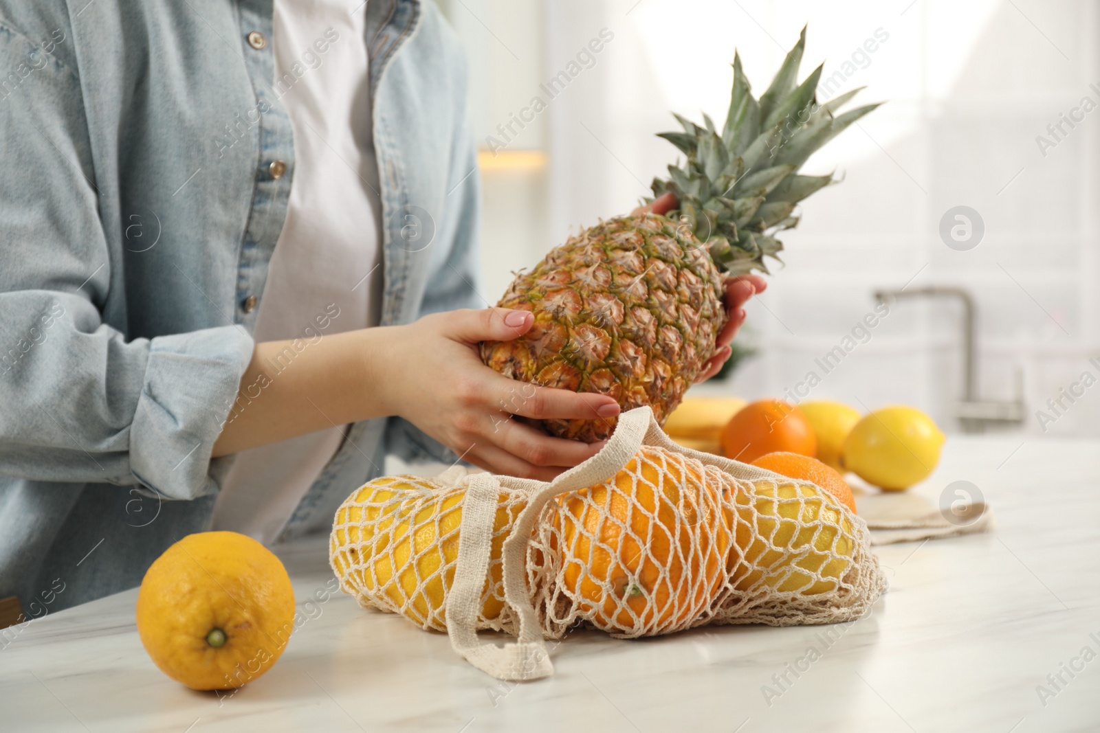 Photo of Woman taking pineapple out from string bag at light marble table, closeup