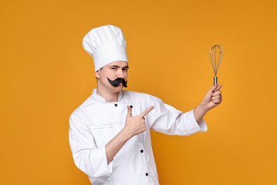 Photo of Portrait of happy confectioner with funny artificial moustache holding whisk on orange background