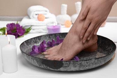 Photo of Woman soaking her feet in bowl with water and flowers on white wooden floor, closeup. Spa treatment
