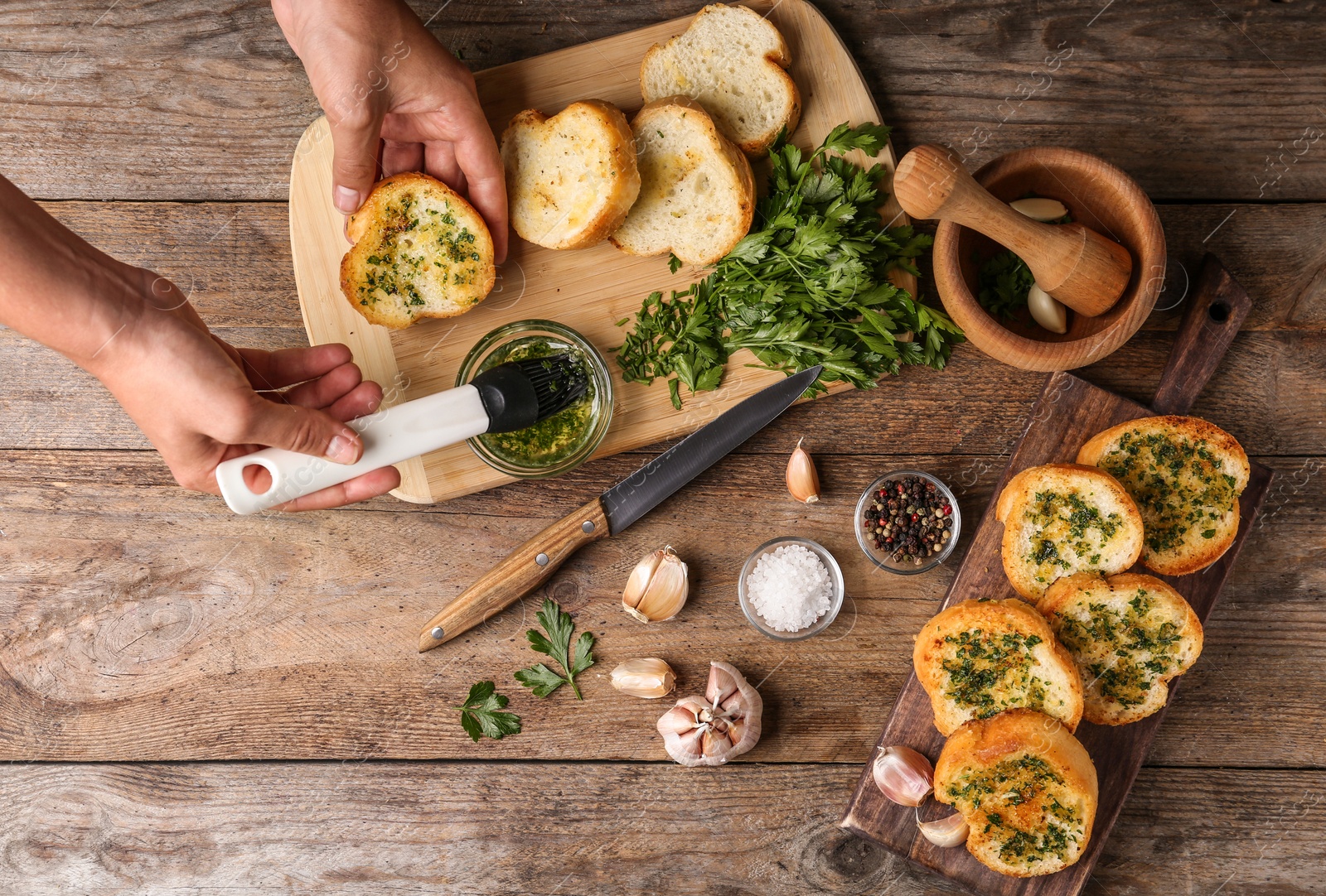 Photo of Woman brushing slice of toasted bread with garlic and herbs over wooden table, top view