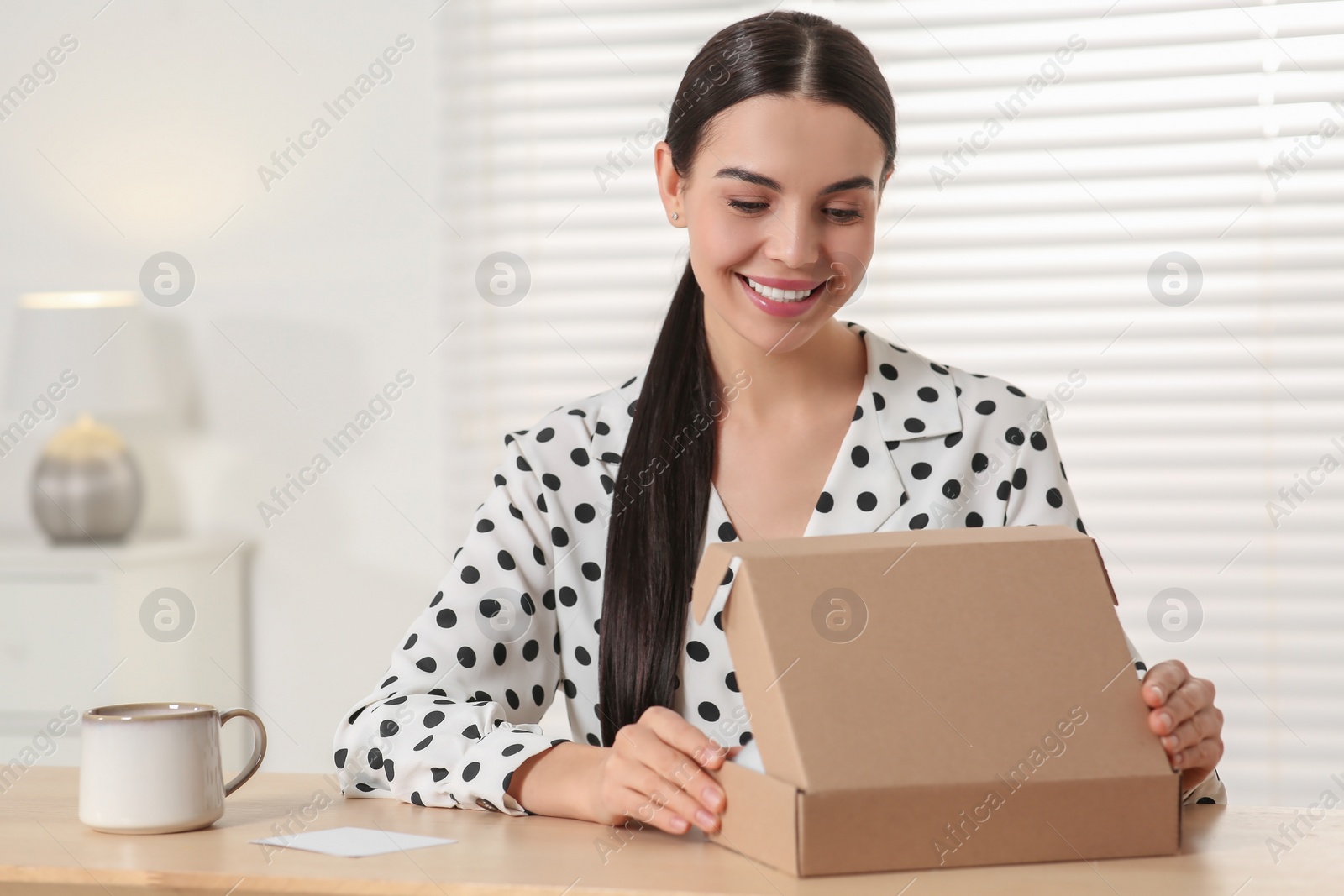 Photo of Happy young woman opening parcel at table indoors. Internet shopping