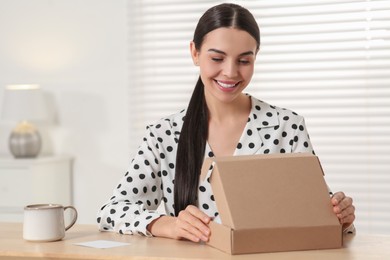 Happy young woman opening parcel at table indoors. Internet shopping