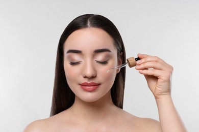 Young woman applying essential oil onto face on white background, closeup
