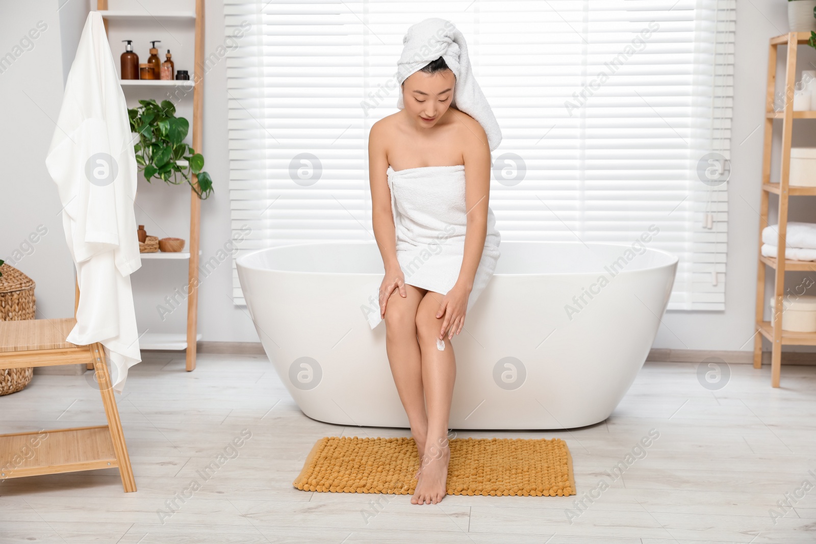 Photo of Beautiful young Asian woman applying body cream on leg in bathroom