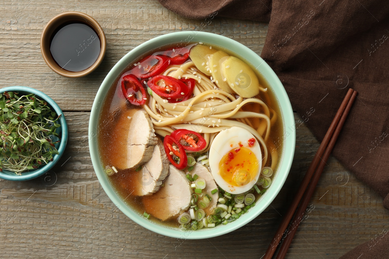 Photo of Delicious ramen in bowl, soy sauce and microgreens on wooden table, flat lay. Noodle soup