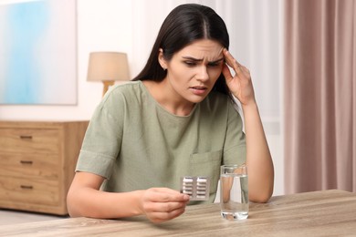 Sad woman with blister of pills suffering from headache at wooden table indoors