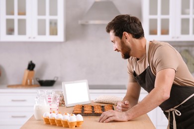 Photo of Man with freshly baked cookies watching online cooking course via tablet in kitchen